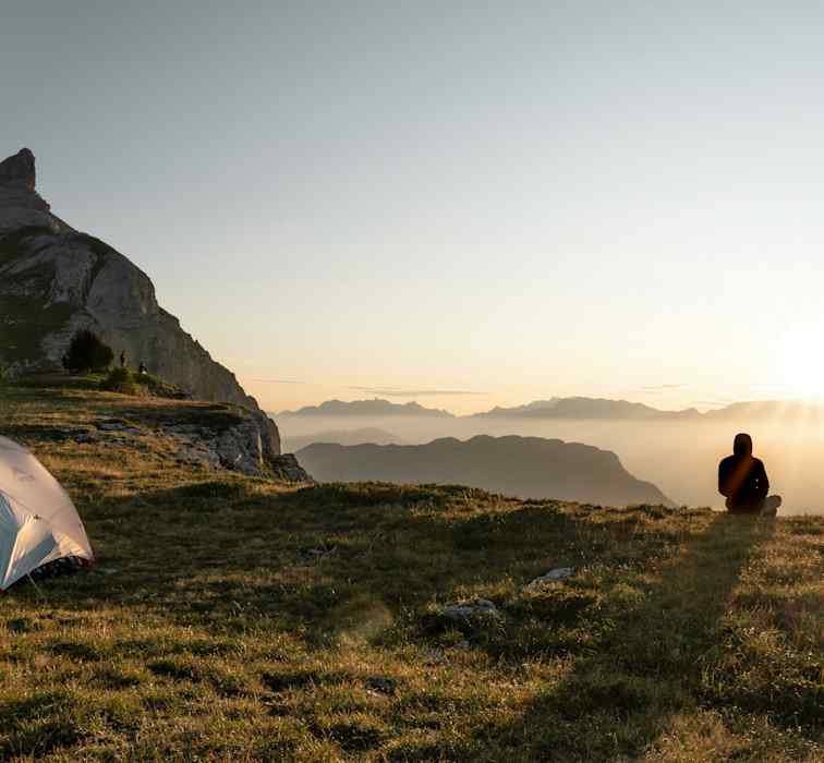 Zwei Personen sitzen vor ihrem Zelt und betrachten den Sonnenuntergang vor einer beleuchteten Bergkulisse.