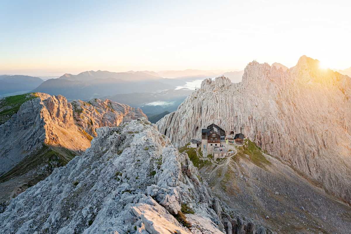Vogelperspektive einer Berghütte, die in spektakulärer Lage in felsiger Landschaft steht, im Hintergrund geht die Sonne unter