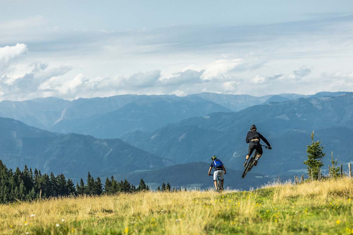 Zwei Mountainbiker fahren über eine Wiese, einer der beiden macht einen Sprung; im Hintergrund die Berge