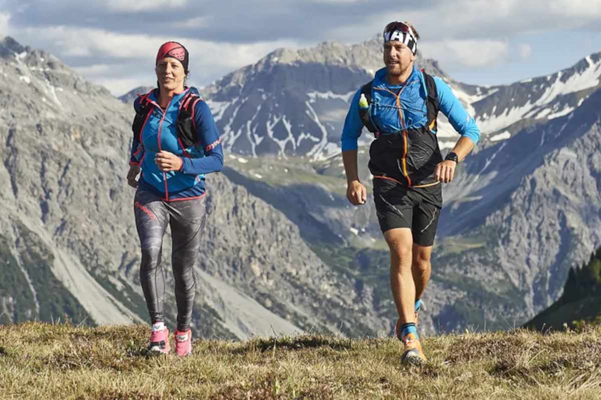 Zwei Trailrunner laufen über eine grüne Wiese. Im Hintergrund eine graue Berglandschaft mit weißen Schneeresten.