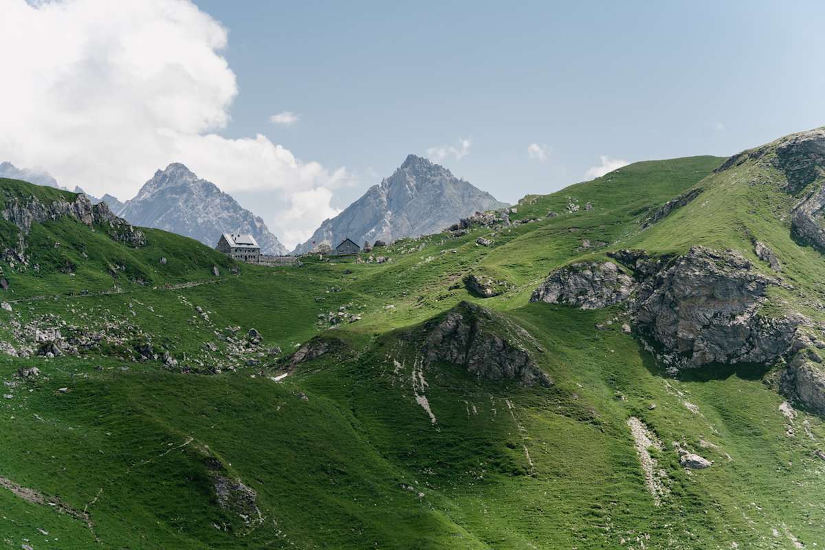 Berglandschaft mit grünen Wiesen, auf einem Kamm in der Ferne steht eine Berghütte aus Stein