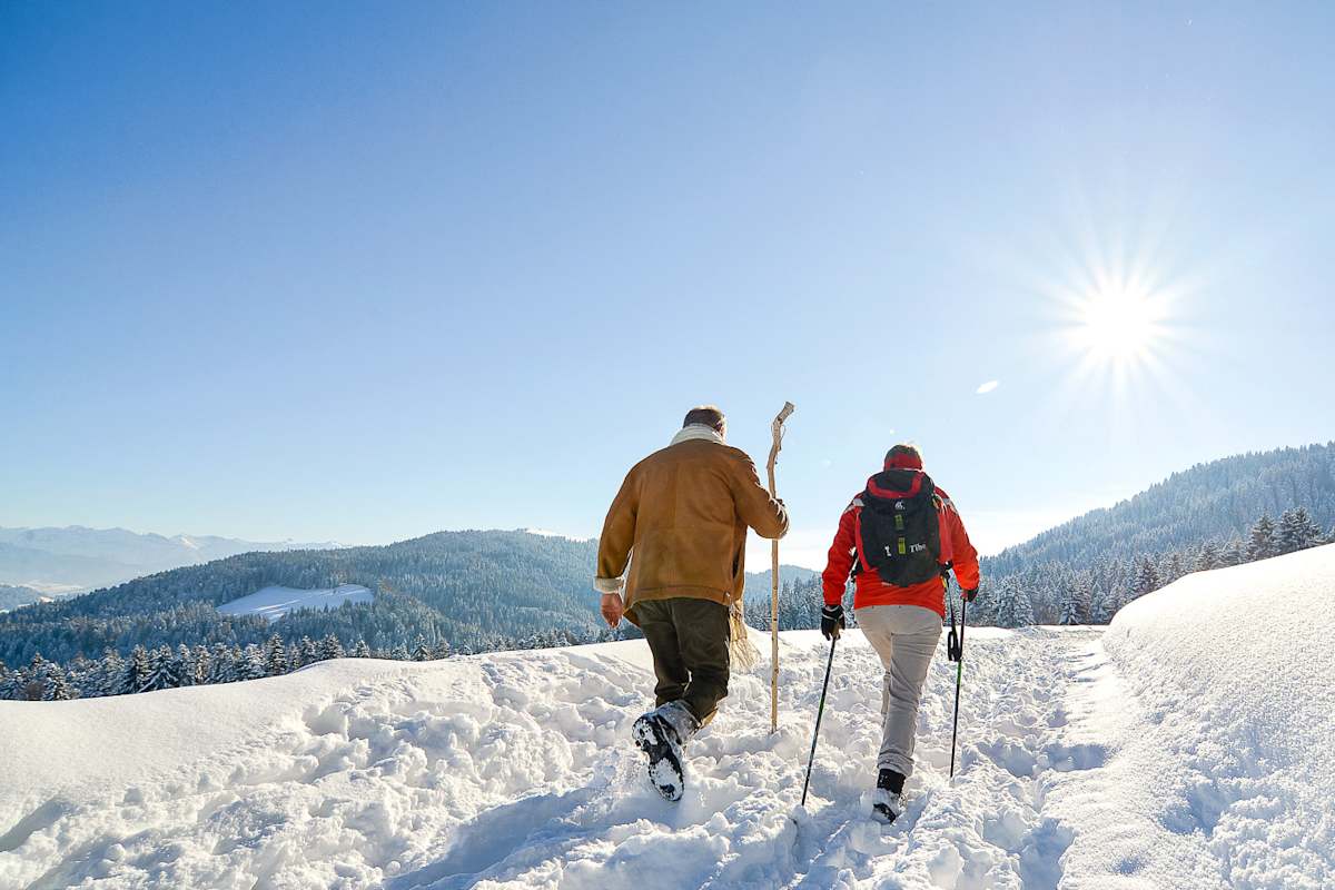 Zwei Winterwanderer stapfen in Scheidegg durch knöcheltiefen Schnee. Im Hintergrund verschneite Wälder und Berge.