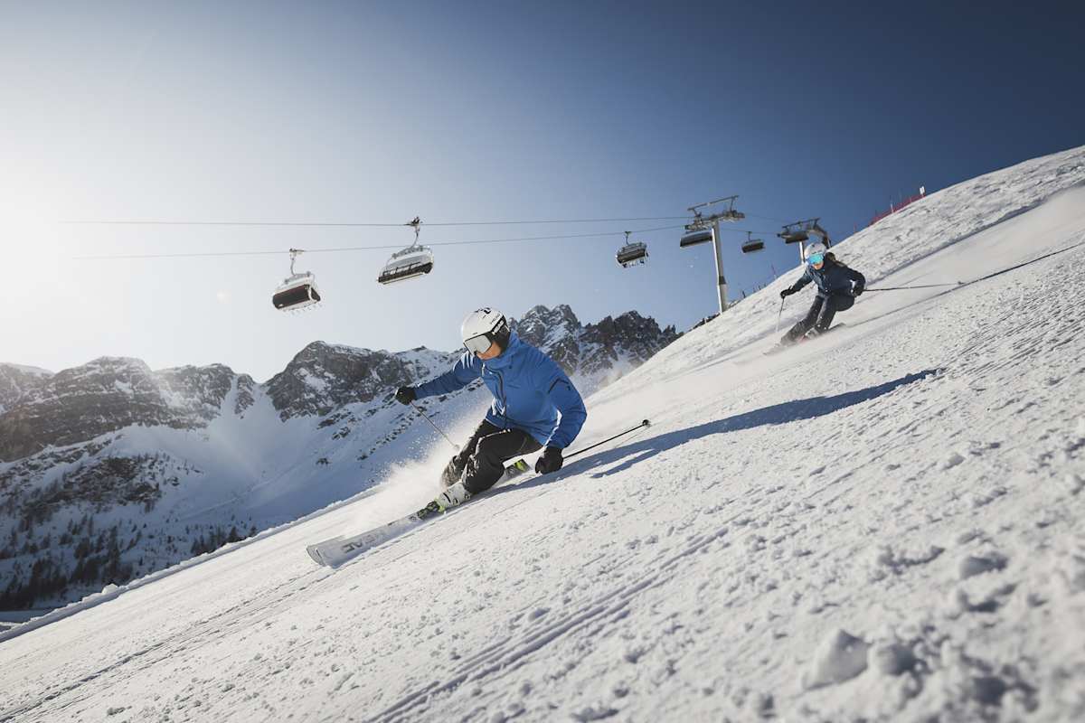 Ein Skifahrer und eine Skifahrerin fahren eine präperierte Piste im Skigebiet Ladurns hinab. Hinter ihnen sieht man einen Sessellift.