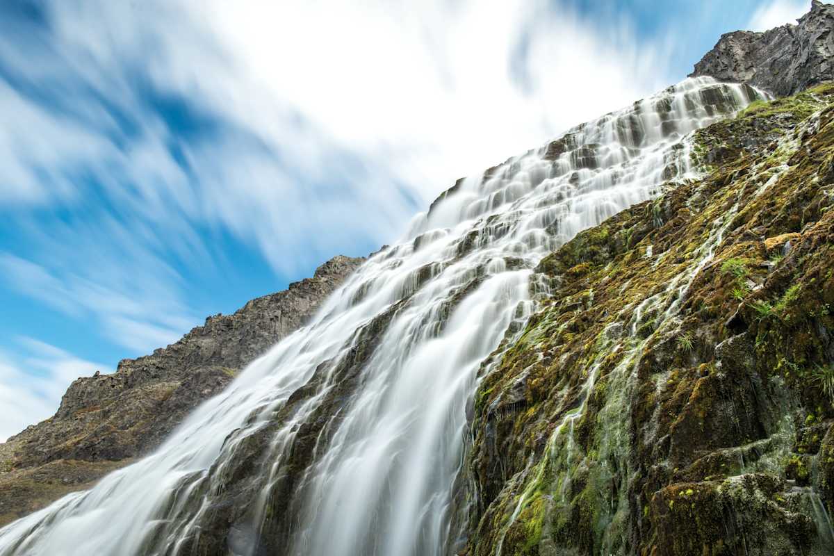 Wasserfall über bemoostem Fels vor leicht bewölktem Himmel