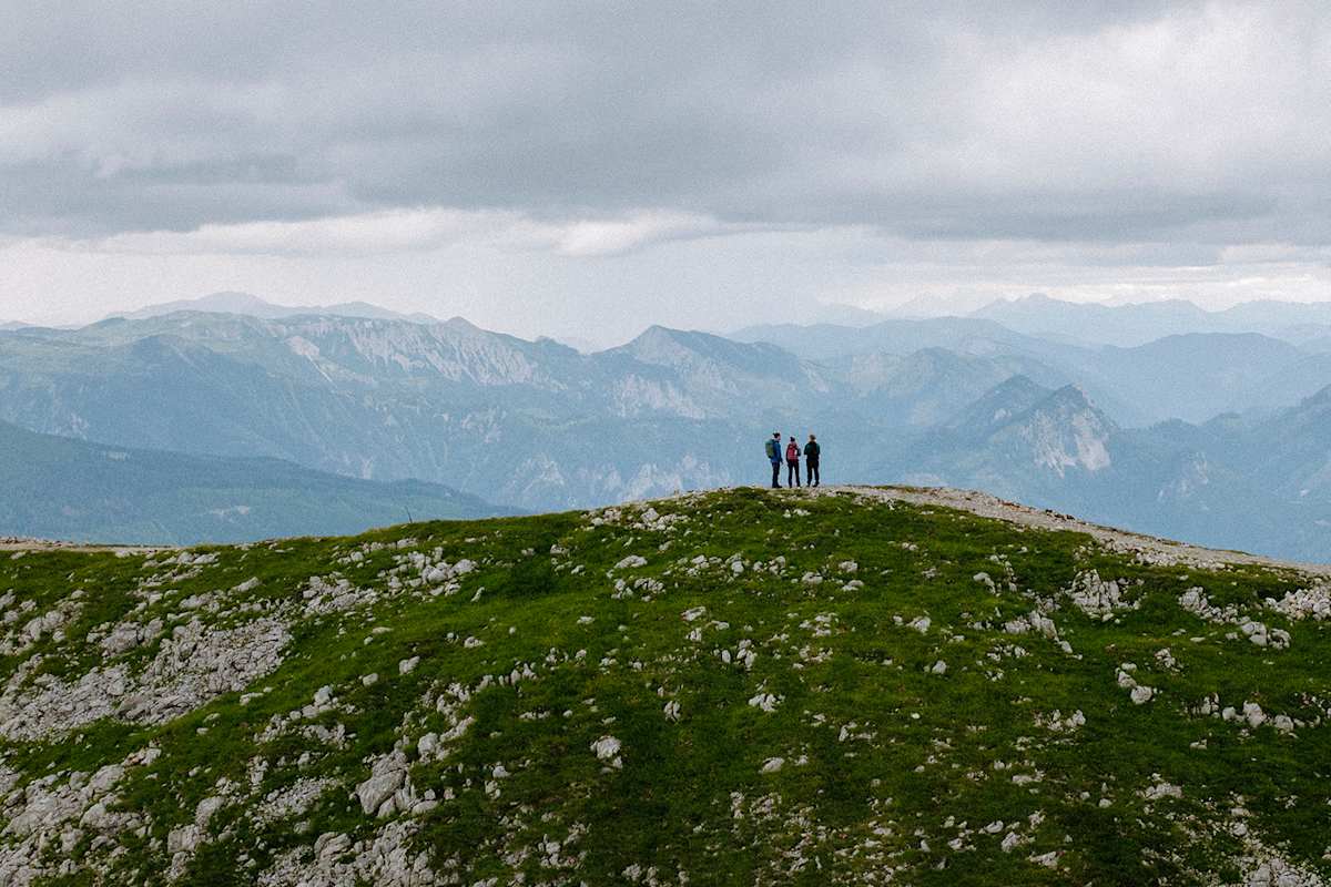 Drei Personen spazieren einen steinigen Berg entlang und blicken auf das bergige Panorama hinab.
