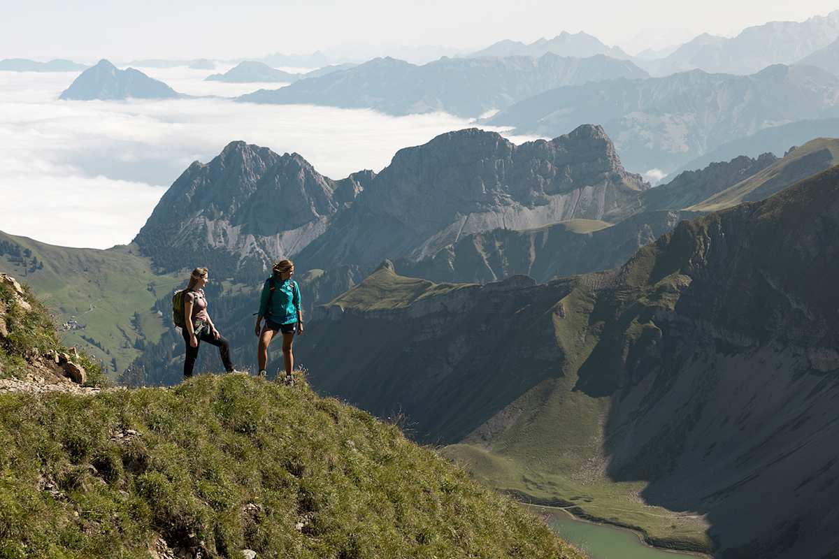 Zwei Frauen stehen auf einer grün bewachsenen Bergspitze und schauen zu den gegenüberliegenden Gipfeln hinüber.