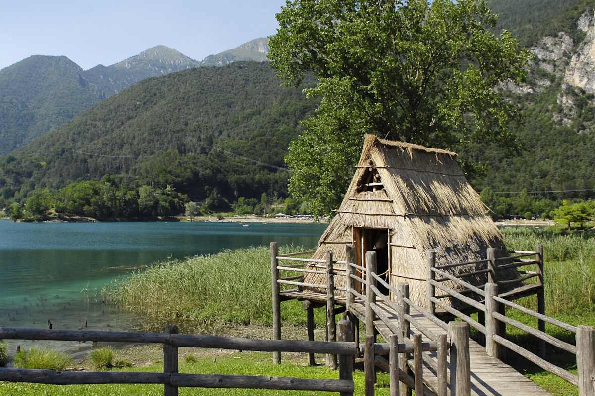 Ein strohgedeckter Pfahlbau an einem grünen See.