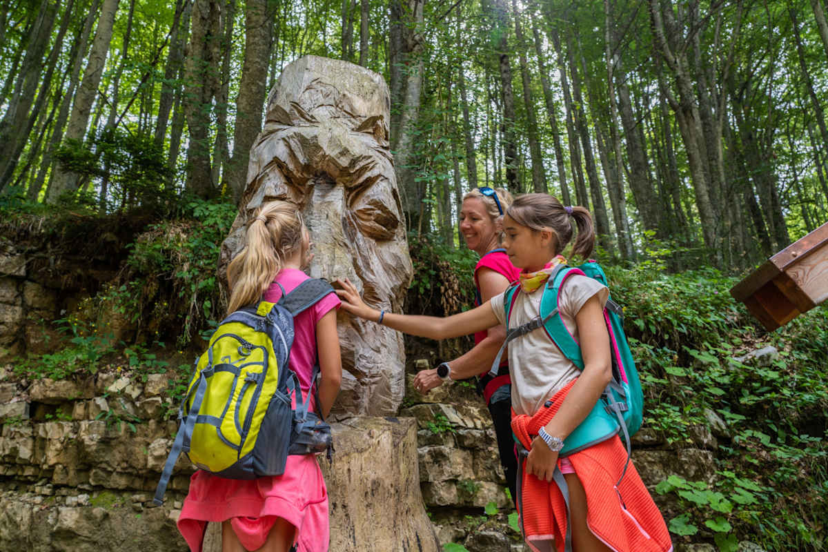 Ein Mutter mit zwei Kindern bewundert eine Holzfigur auf dem Phantasiepfad auf der Alpe Cimbra.