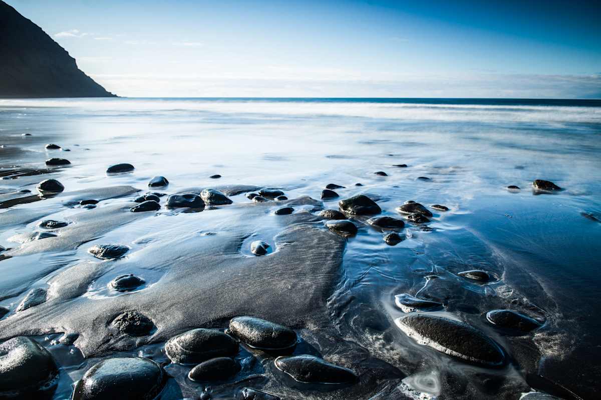 Dunkler Sand mit nassen Steinen bei Ebbe und Meer im Hintergrund