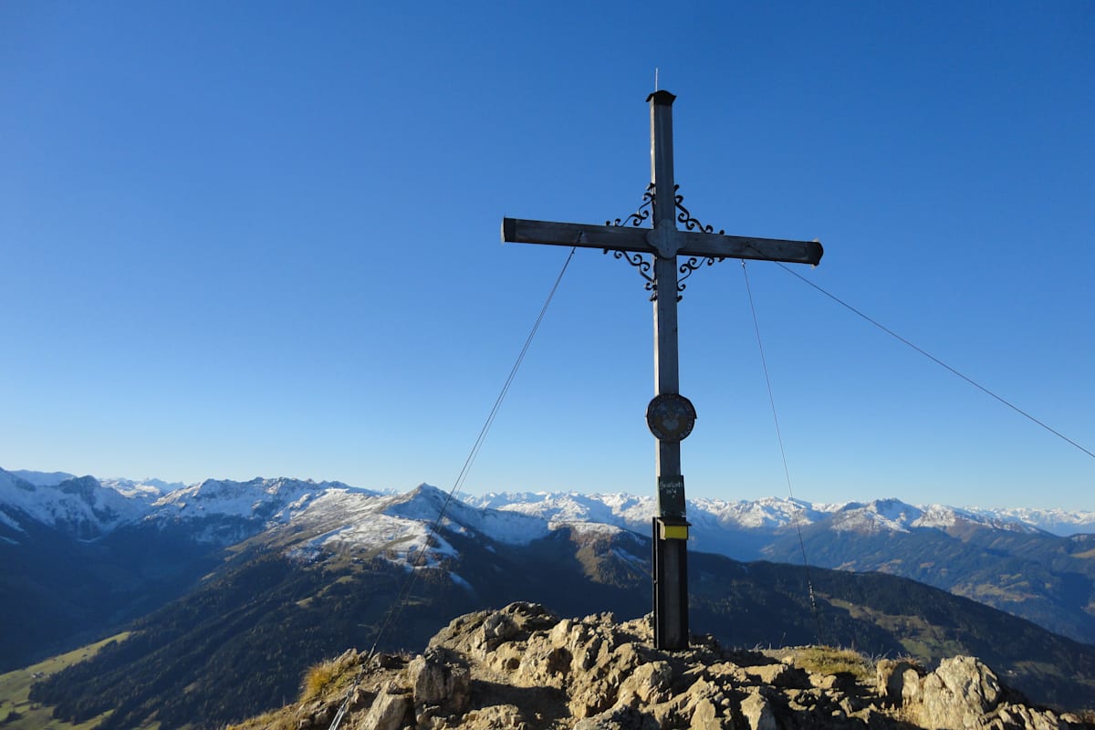 Zu sehen ist ein Gipfelkreuz mit einem sonnigen, alpinen Panorama im Hintergrund.