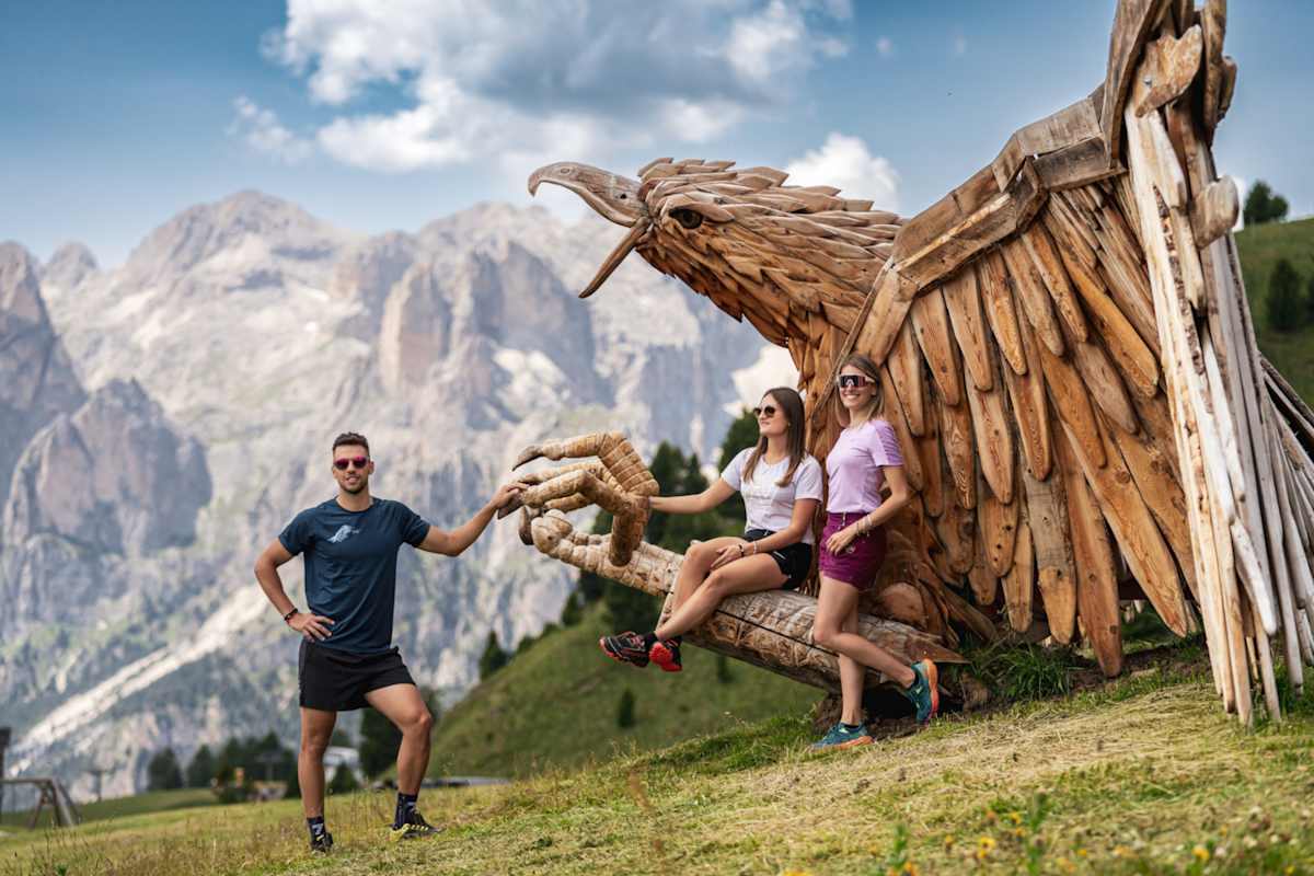 Drei Personen stehen vor einem großen Holzadler. Im Hintergrund die markanten Gipfel der Dolomiten.