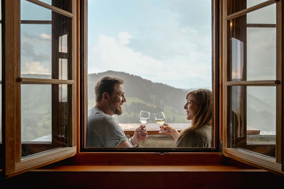 Ein Paar genießt ein Glas Wein mit Blick auf die Berglandschaft in Gstaad durch ein offenes Fenster.