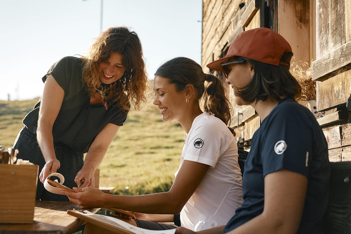 Zwei Frauen sitzen auf einer Bank bei der Einkehr in einer Gasthütte und lassen sich von der freundlichen Kellnerin die Karte erklären.