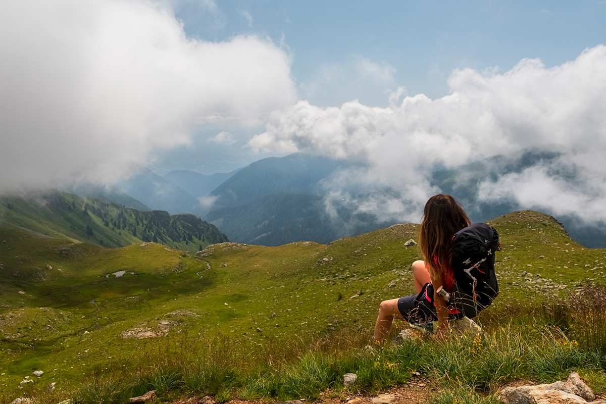 Eine Wanderin rastet auf einer grünen Hochgebirgswiese und blickt durch die Wolkendecke ins Tal.
