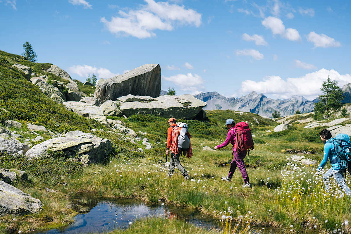 Drei Personen gehen mit Kletterseilen ausgerüstet hintereinander in einer malerischen, bergigen Landschaft.
