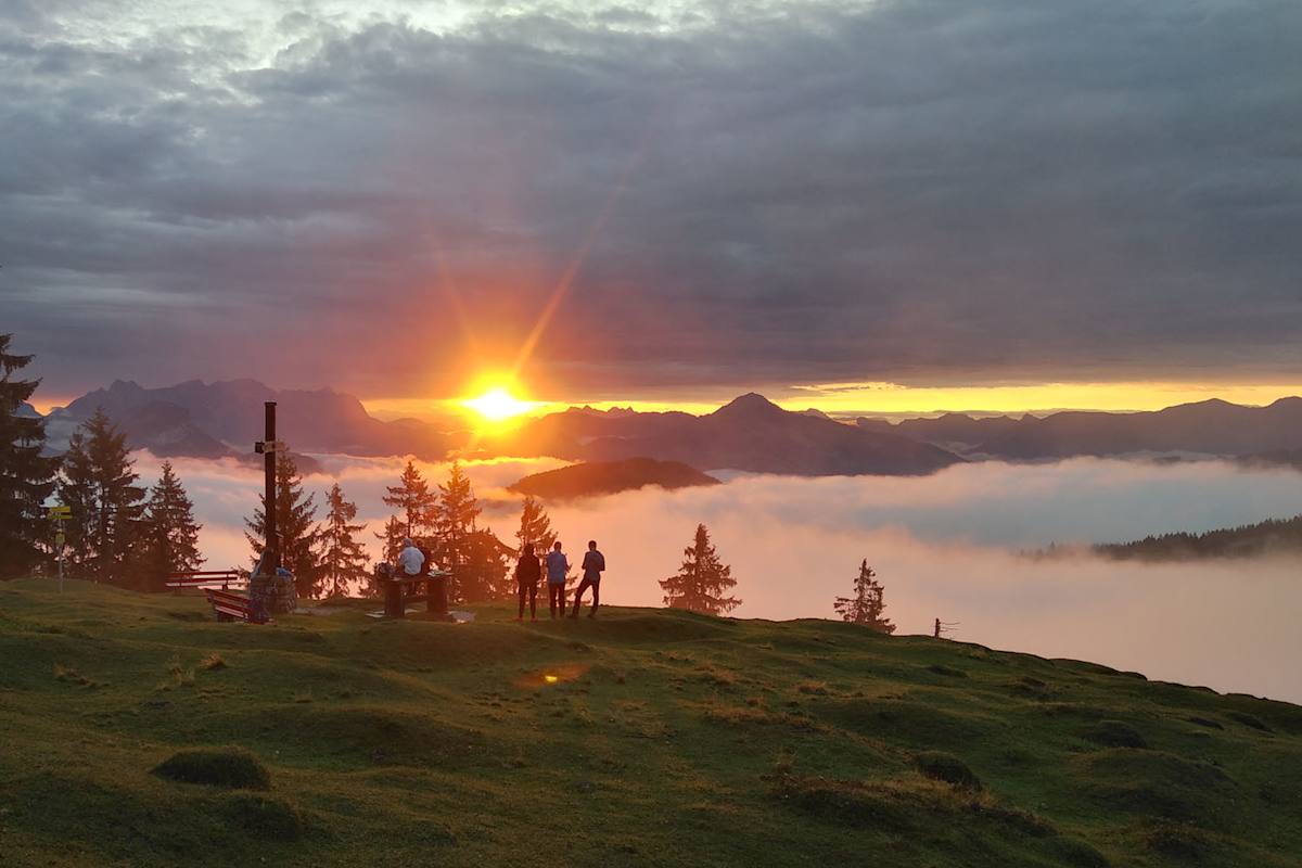 Man sieht die kleinen Silhouetten mehrerer Wanderer die sich auf einer Almwiese befinden und den Sonnenuntergang über den gegenüberliegenden Gipfeln bestaunen.