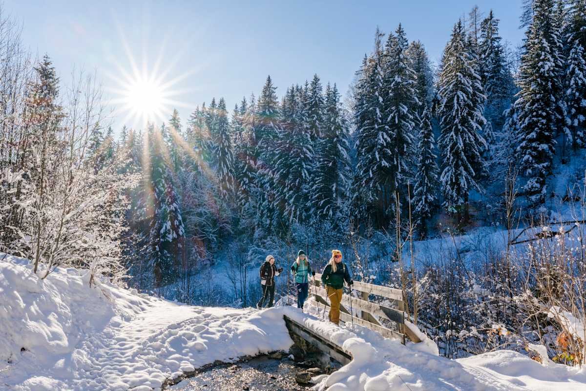 Drei Winterwanderer stapfen durch eine tief verschneite Schneelandschaft am Wilden Kaiser. Sie überqueren gerade eine eingeschneite Brücke über einen kleinen Bach.