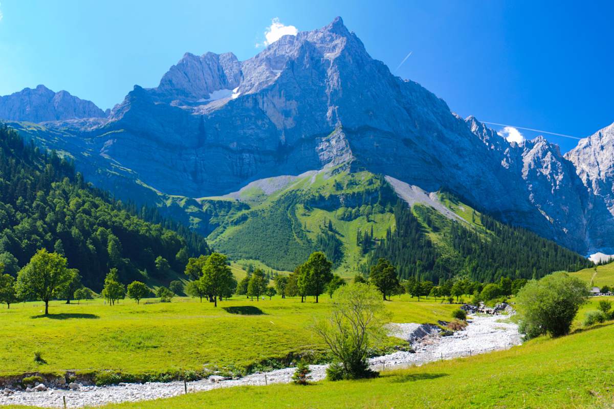 Ein schroffer Berggifpel, davor eine grüne Wiese mit ausgetrocknetem Bach.