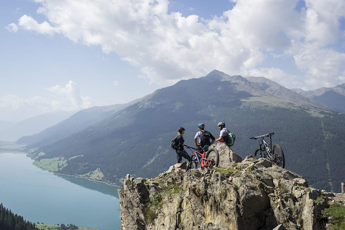 Drei Biker machen Pause auf der Bergspitze mit Blick auf einen See.