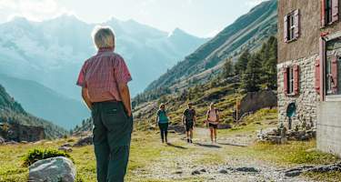 Ein Mann steht vor einer Hütte und wartet auf zwei Wanderinnen und einen Wanderer.