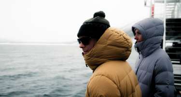 Zwei Personen stehen in Daunenjacken auf einem Schiff - hinter ihnen das Meer
