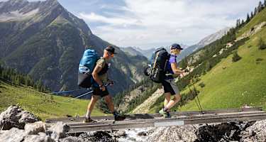 Zwei Wanderer in den Lechtaler Alpen gehen über einen schmalen Holzsteg über einen Bach; im Hintergrund sieht man die Bergwelt