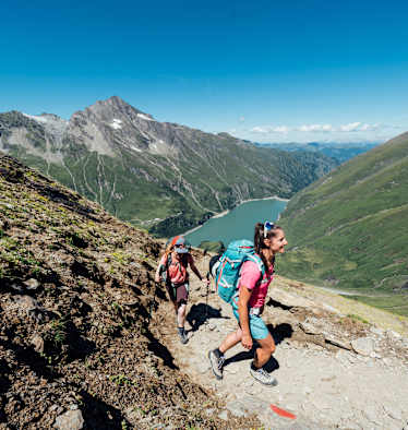 Zwei Frauen Wandern in der Sonne - im Hintergrund ein Stausee und die Berge