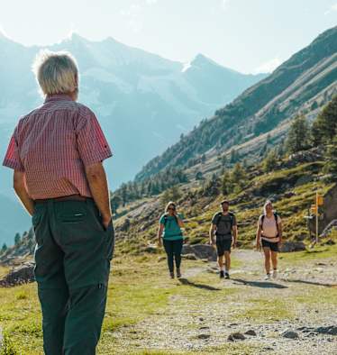 Ein Mann steht vor einer Hütte und wartet auf zwei Wanderinnen und einen Wanderer.
