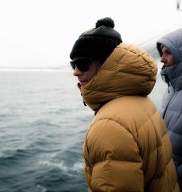 Zwei Personen stehen in Daunenjacken auf einem Schiff - hinter ihnen das Meer