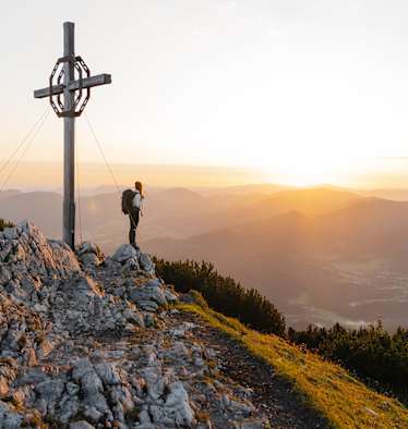 Eine Dame steht neben einem Gipfelkreuz auf einem Felsen und sieht in ein sonniges Tal hinab.