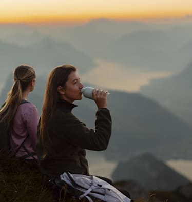 Zu sehen sind zwei Frauen, die auf der Spitze eines Berges sitzen und das wunderbare spätabendliche Panorama bewundern.