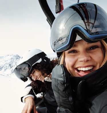 Eine Frau mit Skibrille und Helm auf dem Kopf sitz in einem Sessellift und lacht uns entgegen. Neben ihr sitzt eine weitere lächelnde Frau.