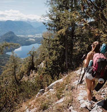 Eine Wandrerin mit Rucksack und Ausrüstung, die durch den Wald auf den Altauseer See hinunterblickt.
