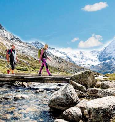 Wanderer überqueren einen Gebirgsbach, schneebedeckte Berge im Hintergrund