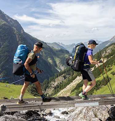 Zwei Wanderer in den Lechtaler Alpen gehen über einen schmalen Holzsteg über einen Bach; im Hintergrund sieht man die Bergwelt
