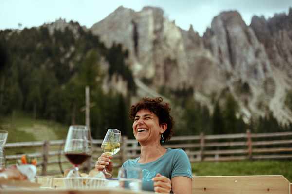 Frau auf einer Alm lacht und hält ein Glas Wein in der Hand - im Hintergrund das Latemarmassiv im Eggental.