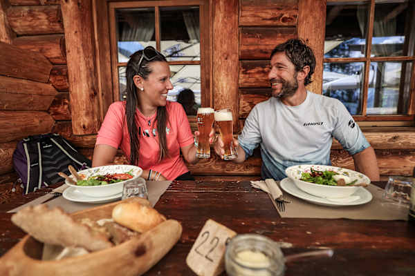 Latemar Ronda, Eggental: Zwei Personen auf der Alm stoßen mit Bier an und haben volle Teller vor sich.