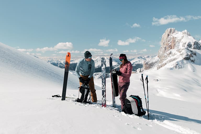 Eine Frau und ein Mann stehen im Schnee auf einem Berg und machen sich für das Skifahren bereit.