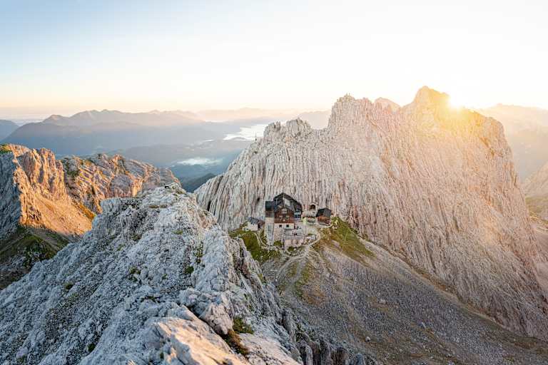 Vogelperspektive einer Berghütte, die in spektakulärer Lage in felsiger Landschaft steht, im Hintergrund geht die Sonne unter