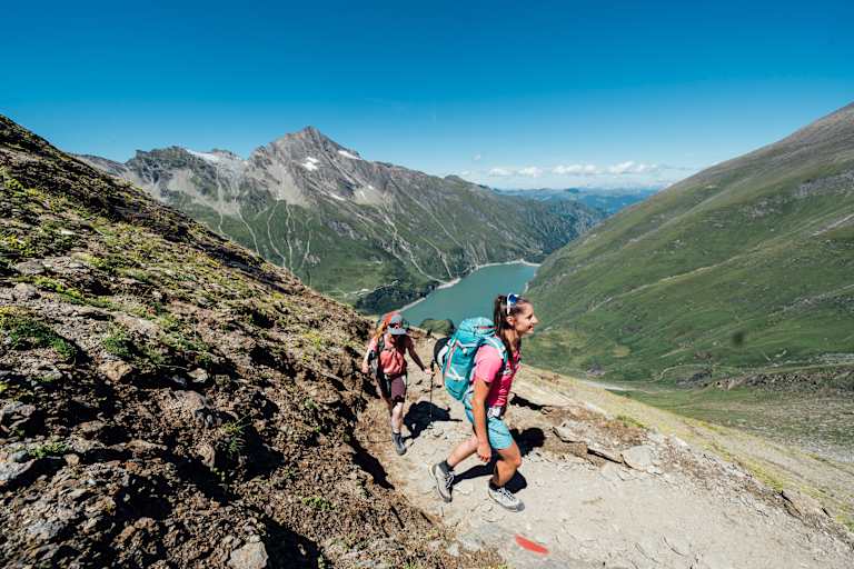 Zwei Frauen Wandern in der Sonne - im Hintergrund ein Stausee und die Berge