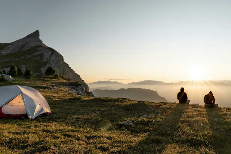 Zwei Personen sitzen vor ihrem Zelt und betrachten den Sonnenuntergang vor einer beleuchteten Bergkulisse.