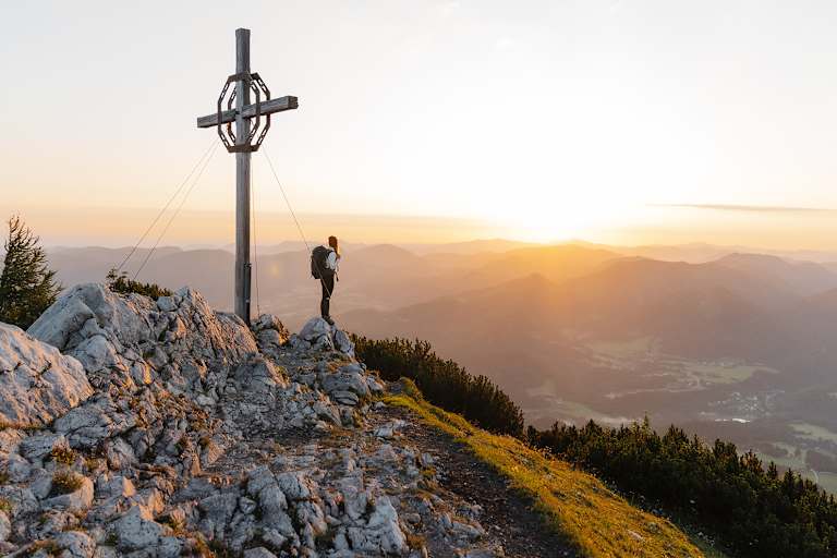 Eine Dame steht neben einem Gipfelkreuz auf einem Felsen und sieht in ein sonniges Tal hinab.