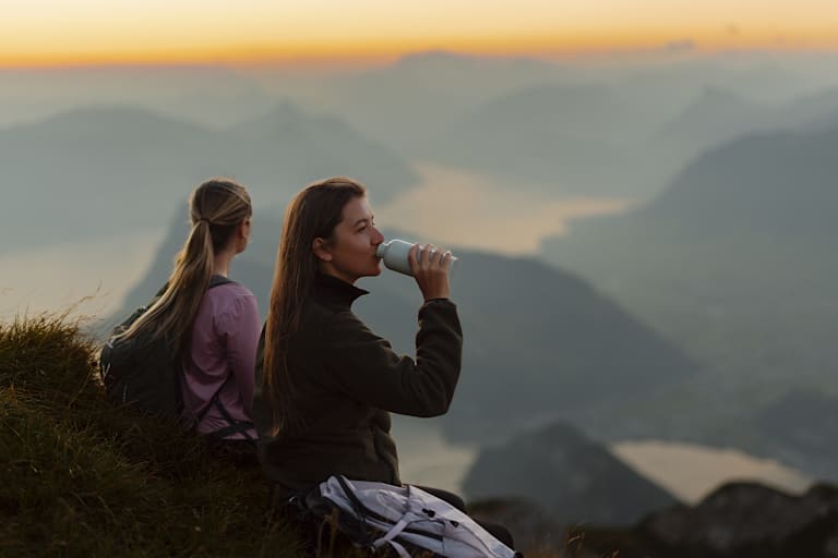 Zu sehen sind zwei Frauen, die auf der Spitze eines Berges sitzen und das wunderbare spätabendliche Panorama bewundern.