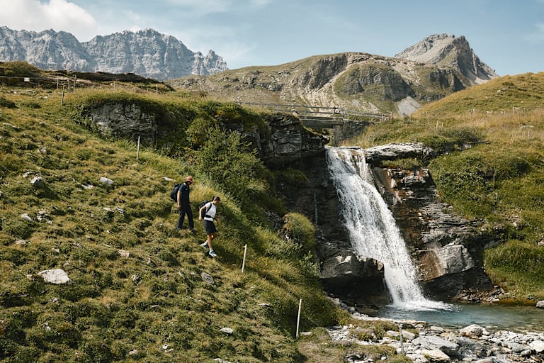 Zwei Frauen wandern einen Hügel hinab. Neben ihnen verläuft ein kleiner Wasserfall und im Hintergrund ist eine alpine Landschaft sichtbar.