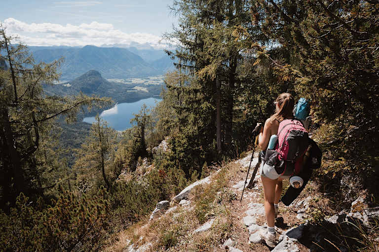 Eine Wandrerin mit Rucksack und Ausrüstung, die durch den Wald auf den Altauseer See hinunterblickt.