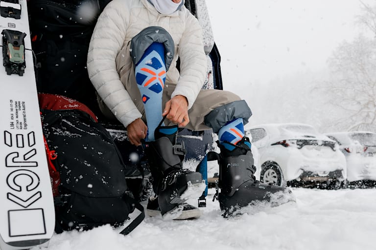 Ein Skifahrer sitzt am Parkplatz und zieht sich im Schnee seine Skischuhe an.