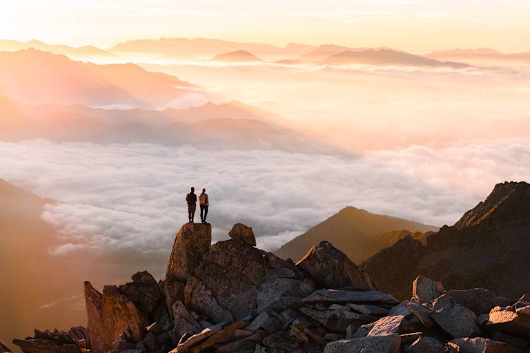 Zwei Wanderer auf einem Felsen im Hochgebirge überblicken das von Wolken bedeckte Tal.