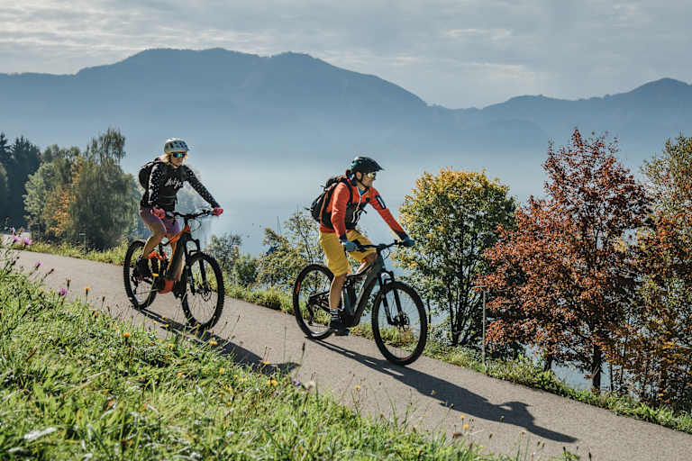 Zwei Radfahrer auf einer Straße in Oberösterreich, im Hintergrund Berge und buntgefärbte Bäume.