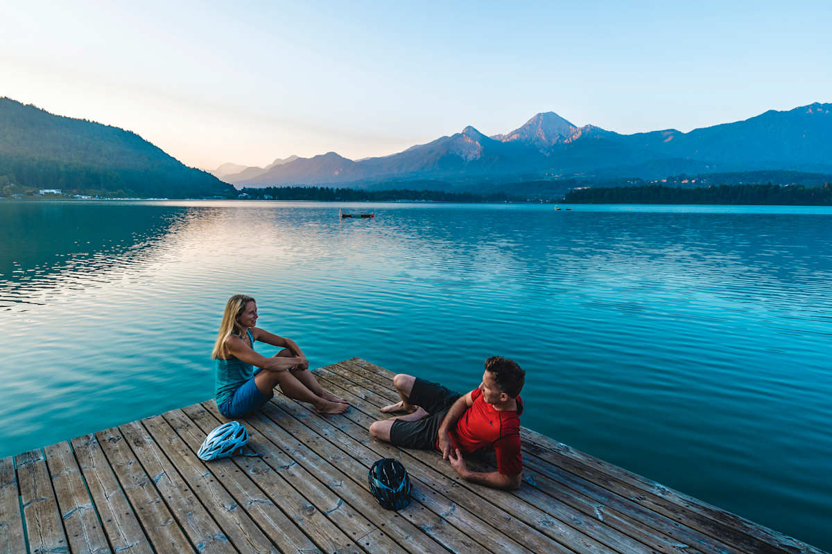 Zwei Radfahrer enstpannen auf einem Holzsteg am Faaker See.