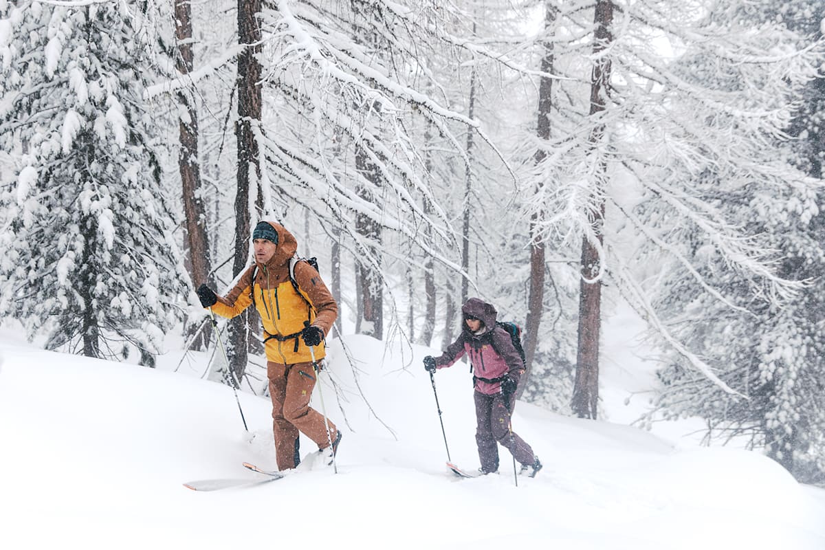 2 Personen gehen mit Ski und Ausrüstung durch den verschneiten Wald im Winter.